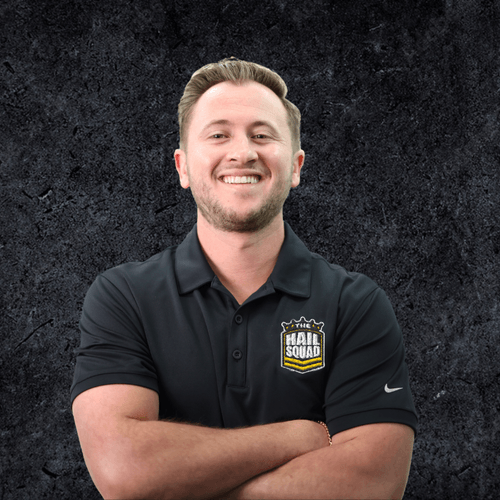 A smiling person with short hair stands confidently in a black polo shirt adorned with "The Hail Squad" logo, set against a dark textured background, exuding a sense of home and belonging.