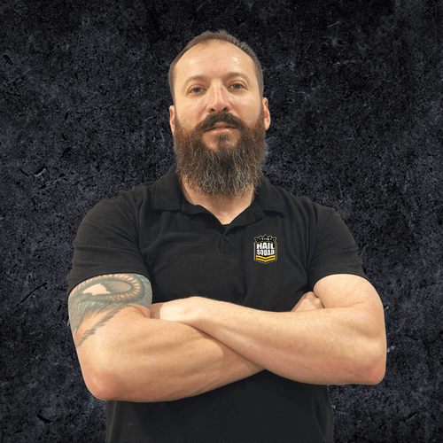 A man with a beard and tattoos stands with arms crossed at home, set against a dark textured background, wearing a black polo shirt.