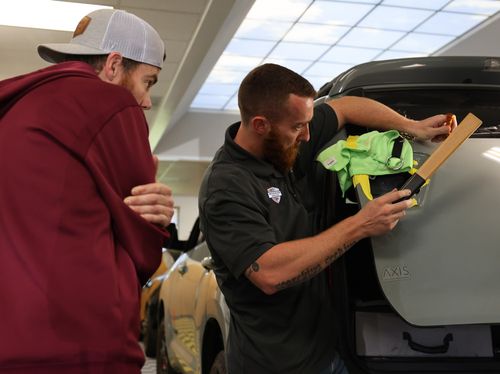 Two men are examining the open trunk of a car indoors, with one holding a measuring tool and safety vests, preparing for some paintless dent repair.