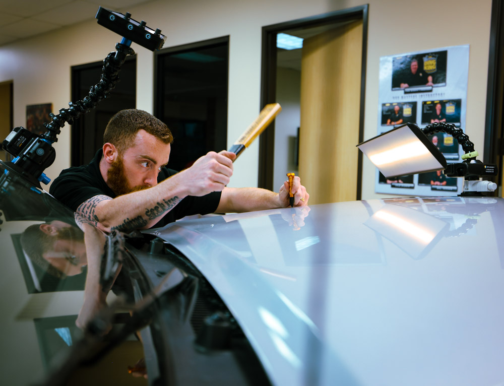 A man expertly performs paintless dent repair on a car hood using a specialized tool in a well-lit workshop.