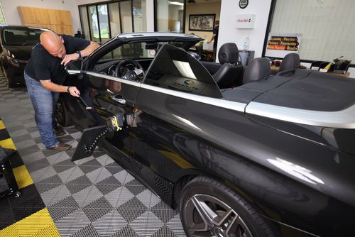 In the garage, a man expertly installs a stereo system in a sleek black convertible car, ensuring every detail complements its pristine finish, just like paintless dent repair would preserve its flawless exterior.