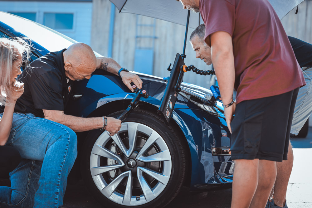 People inspecting the front wheel of a parked blue car, possibly considering paintless dent repair, with an umbrella providing shade.