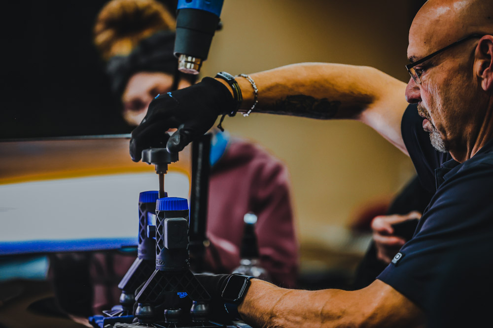 A man wearing gloves operates a machine, tightening a screw for paintless dent repair. Another person stands in the background, observing the precise work.