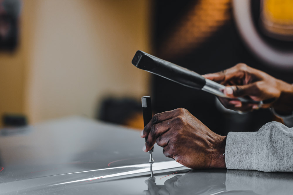 A person is expertly performing paintless dent repair on a car hood, skillfully using a tool to restore its original shape.