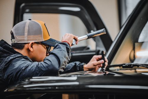 A person in a cap and glasses skillfully repairs a car engine using a tool.