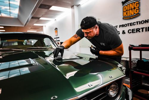 A person wearing a cap and gloves carefully polishes the hood of a green vintage car in a workshop, perfecting every detail while addressing prior hail damage repair.