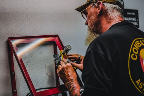 A person with a beard and hat meticulously operates a small tool at a workbench, possibly focusing on hail damage repair as they carefully adjust the red-framed device on the wall.