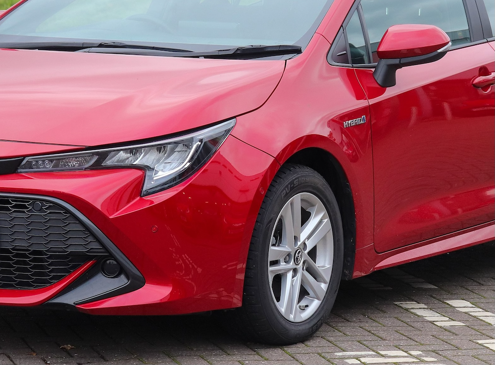 A close-up of the front left side of a red hybrid car parked on the street reveals its headlight, grille, and side mirror, showcasing meticulous hail damage repair that restores its sleek exterior.