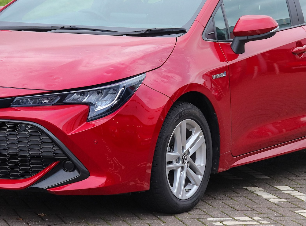 Close-up of a red car parked on a cobblestone surface, showcasing the front-left headlight, grille, and hybrid badge, ready for hail damage repair.