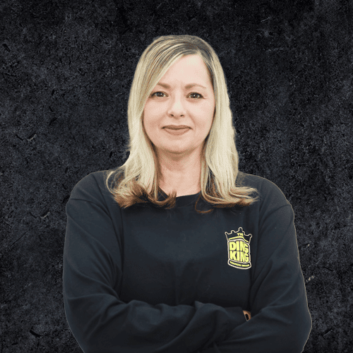 A person with long hair stands against a dark, textured backdrop, wearing a black long-sleeve shirt emblazoned with a yellow home logo on the chest.