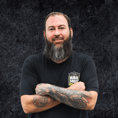 A bearded man with folded arms, wearing a black T-shirt featuring a logo, stands against a textured dark background, exuding a sense of comfort and home.