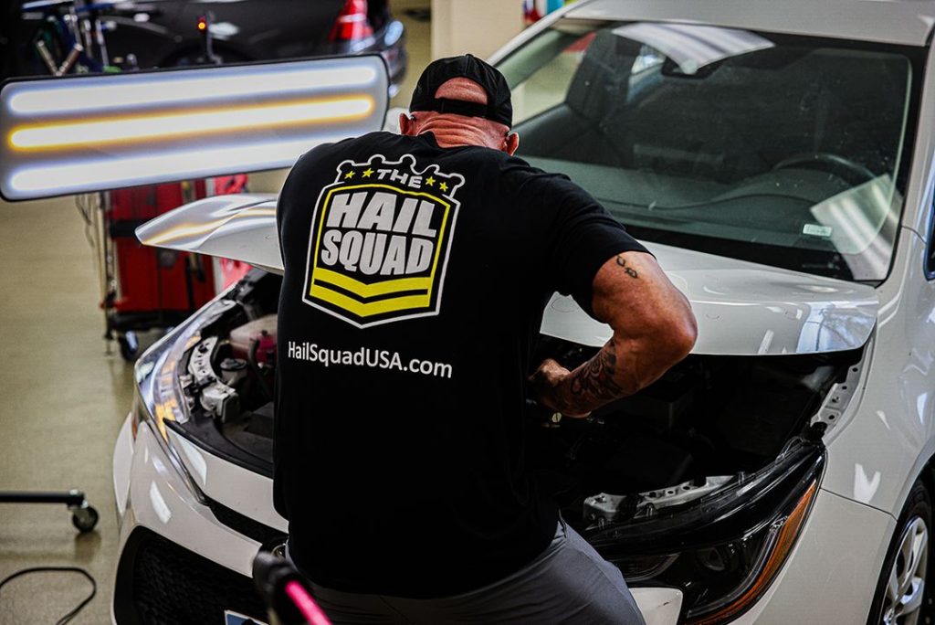 A person sporting a "Hail Squad" t-shirt diligently works on a car in a garage, illuminated by a nearby light panel. The website "HailSquadUSA.com" is prominently displayed, offering insight about The Hail Squad and their passion for automotive excellence.