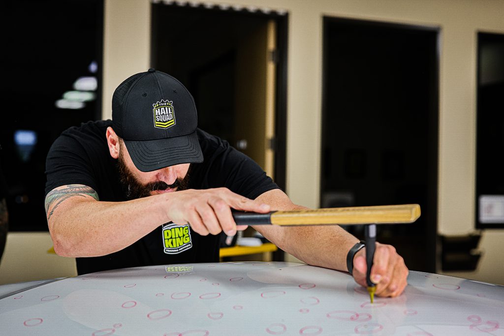 In the workshop, a man in a cap skillfully uses a tool to repair dents on a car's hood, embodying the expertise About The Hail Squad is known for.