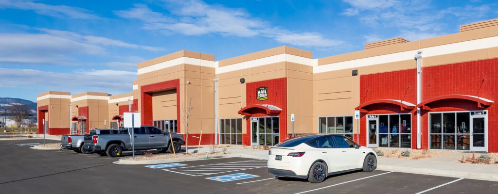 A strip mall with a red and beige facade features multiple storefronts, inviting customers to make contact. Cars are parked in front, under a clear blue sky.