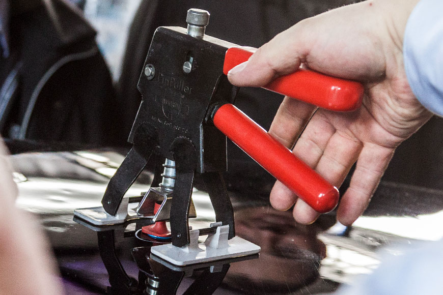 A person from the Hail Squad expertly uses a manual dent puller tool with red handles on a car surface, demonstrating effective paintless dent repair to maintain the vehicle's value.