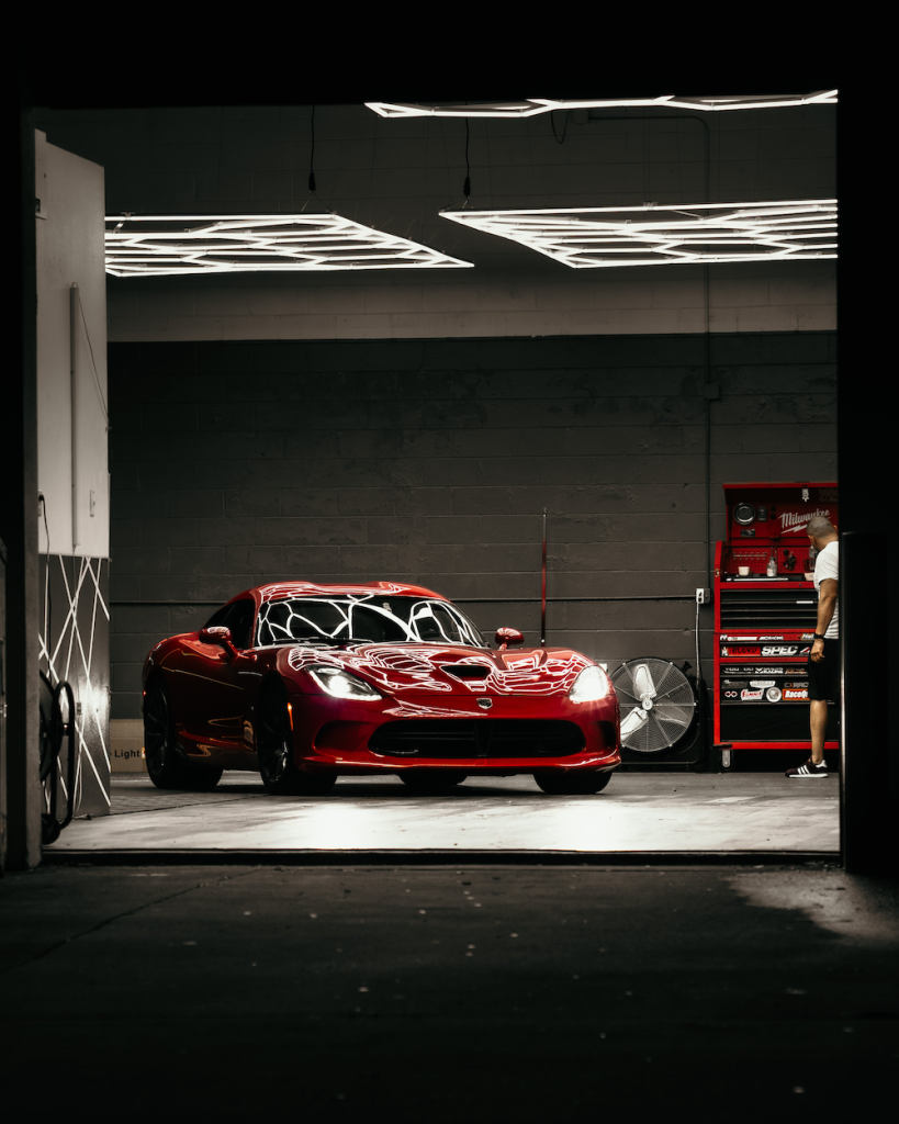 A red sports car gleams in the dimly lit garage, flanked by a tool chest and fan, showcasing its meticulous vehicle care.