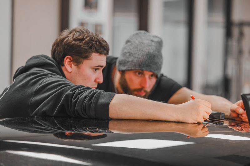 Two men lean over a black surface, intently focused on their task. One wears a beanie and grips a pen; the other sports a hoodie. Their collaboration seems as assured as a lifetime guarantee.