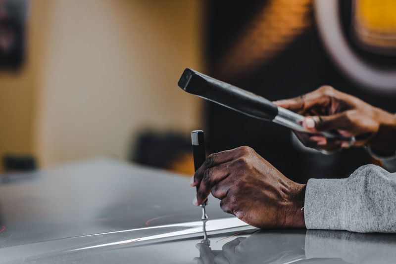 A person skillfully uses tools to repair a dent on the sleek silver surface of a car, ensuring a flawless finish backed by a lifetime guarantee.