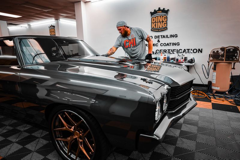 A man with a cap and beard is meticulously polishing the hood of a dark gray vintage car, showcasing the detailing services offered at the shop.