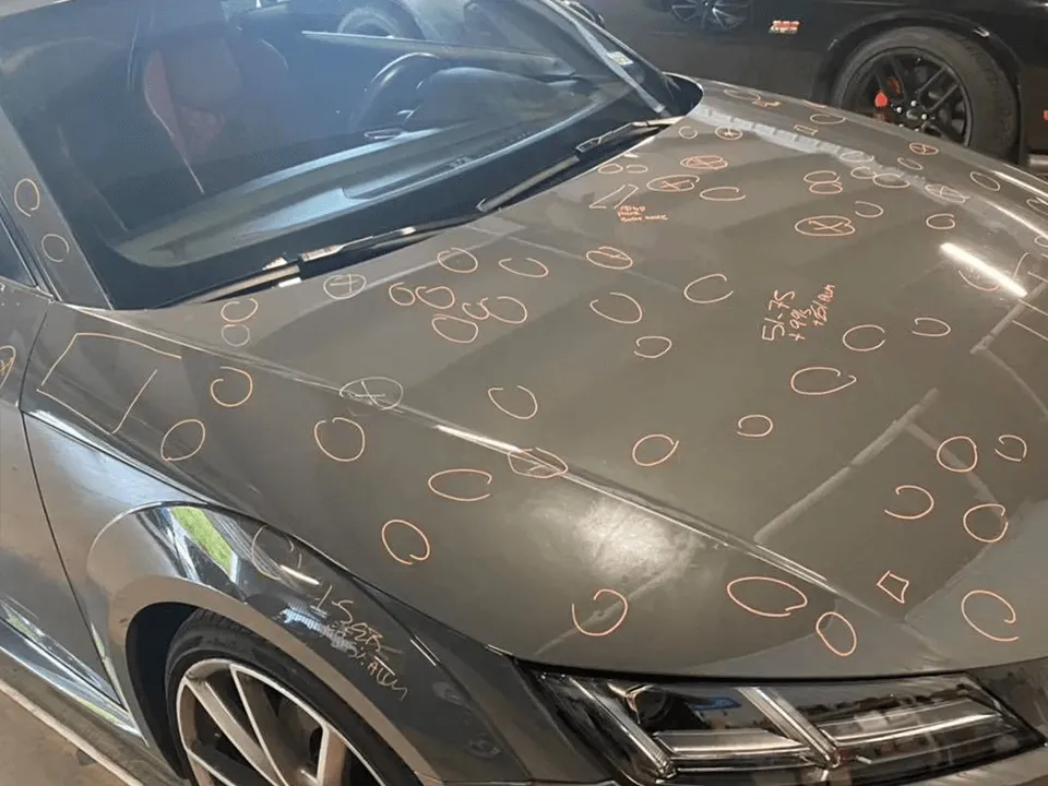 A car sits in the garage, its hood and windshield covered with numerous chalk marks and symbols, indicating areas of vehicle hail damage that were mistakes to avoid during the last storm.